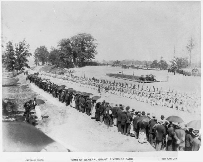 Black and white photo of a crowd of people in a line along grass with a small tomb to the right of them