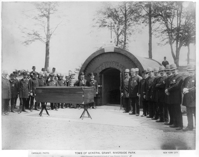 Black and white photo of a coffin placed on a stand, elevated over the ground with a tomb in front and a line of people to the right