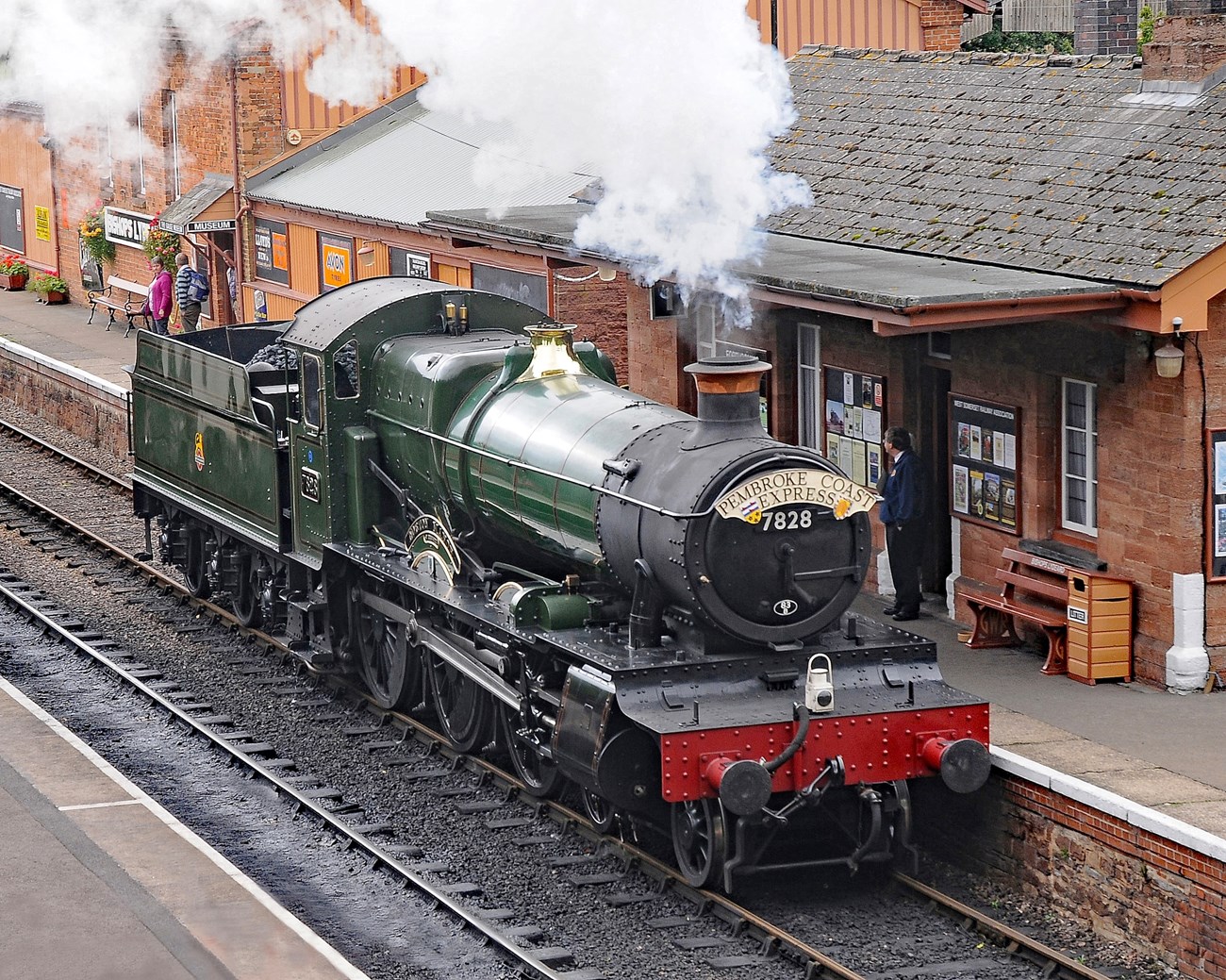 Green and red train with smoke blowing out of it stopping at a train station.