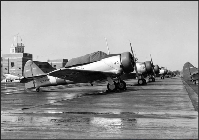 Black and white photo of a line of propeller planes with their cockpits covered on a tarmac. Air control tower is in left background