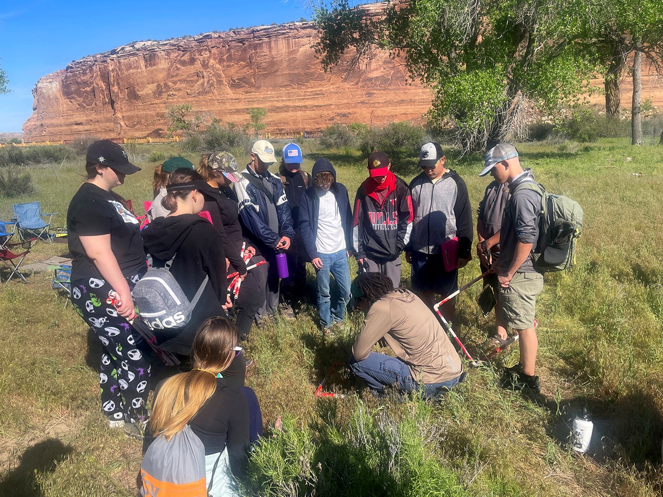 Group of standing students surround a woman sitting on the ground in a grassy field, with measuring equipment. Red rock cliffs surround them.