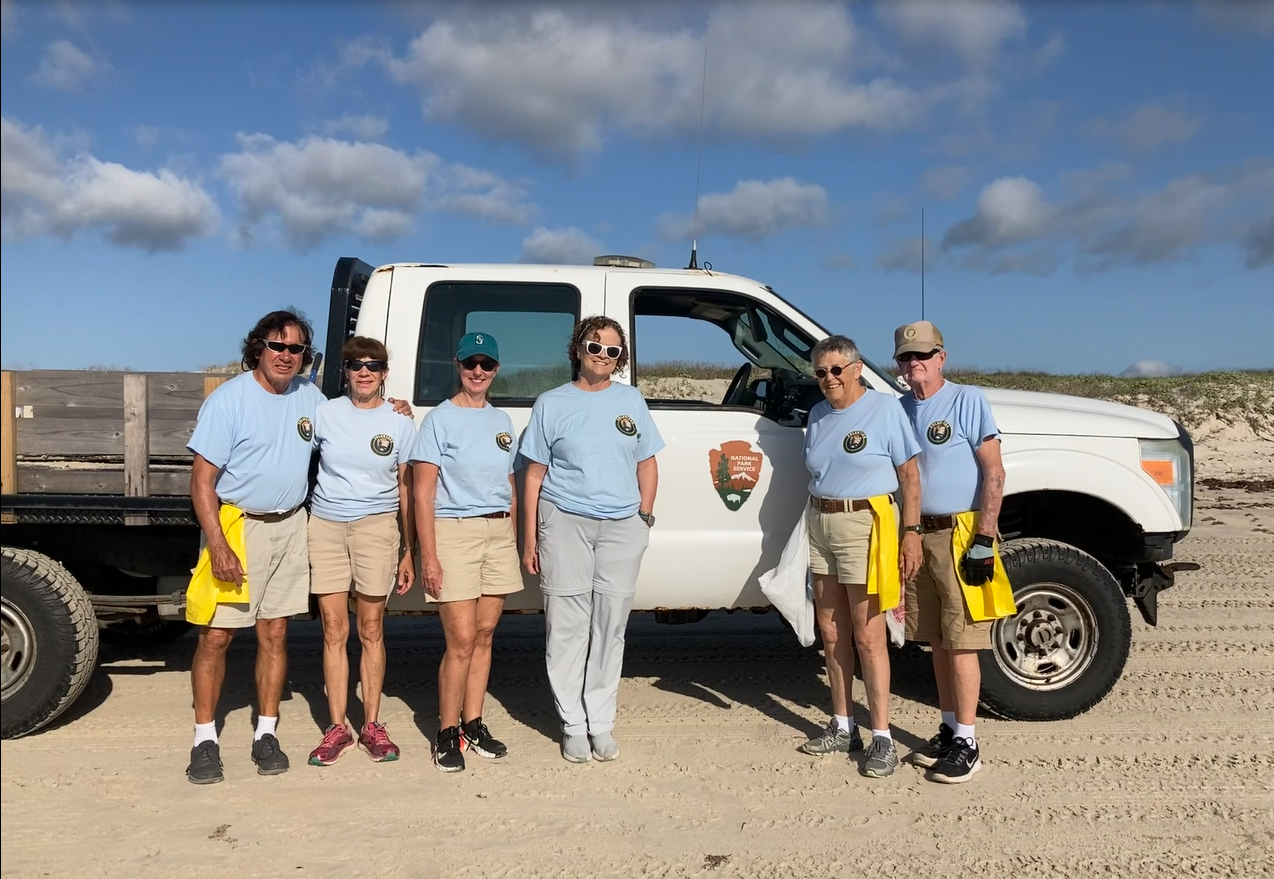 A group of people wearing matching shirts stand next to a white truck parked on a beach.