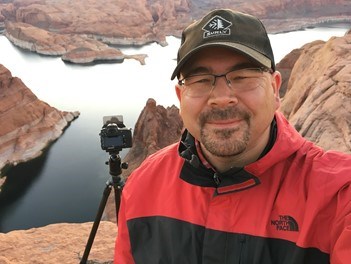 Head and shoulders of a person in front of some rocks and water, with a camera on a tripod.