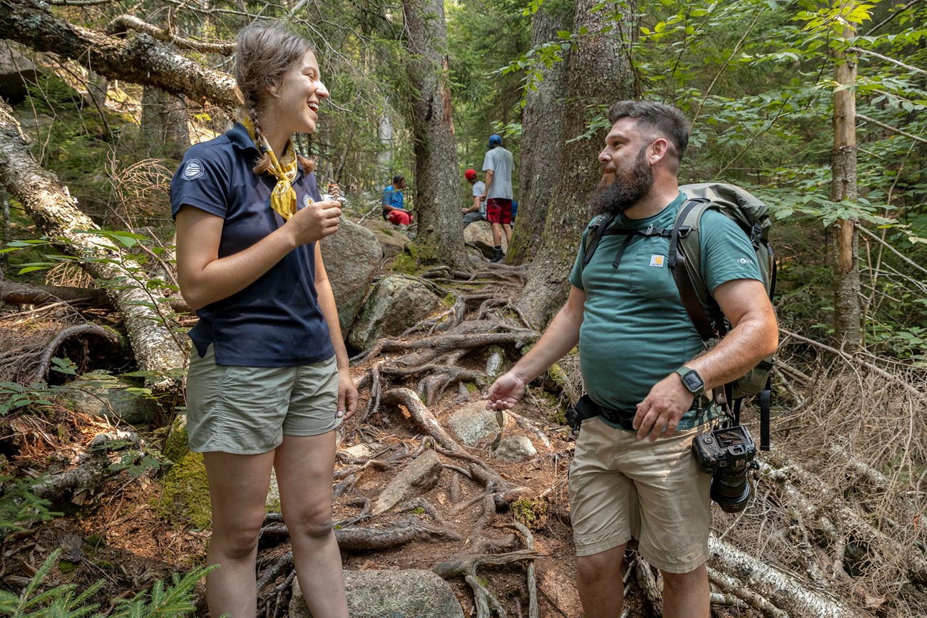 A man with a camera speaking to a woman. They are standing in the woods, with verdant green trees and dark brown earth around them.