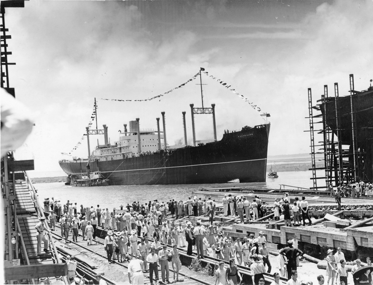 Black and white photo of a crowd gathered on a pier with a large ship festooned with flags just off the dock. Scaffolding on either side