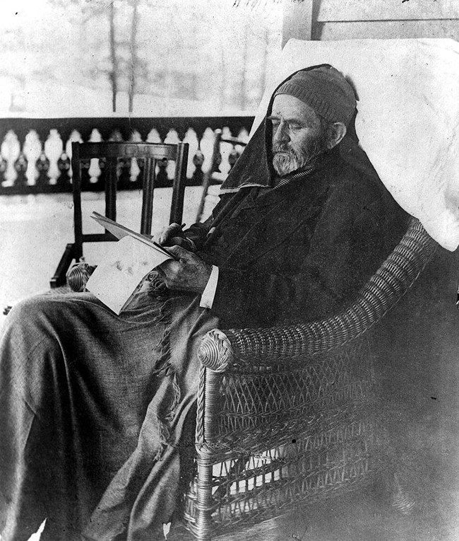 Black and white photo of a man in a warm hat and covered in a blanket in a rocking chair writing in a journal