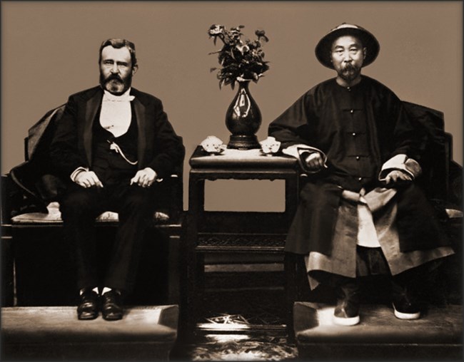 Black and white photo of an American man in a suit on a chair on a raised platform on the left, and a Chinese man on the right in a chair on the same platform