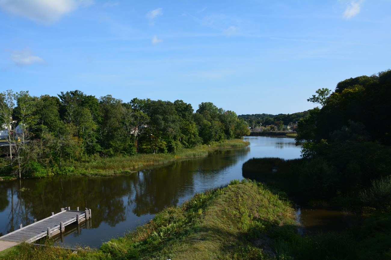 A reflective river under a blue sky between green rows of trees.