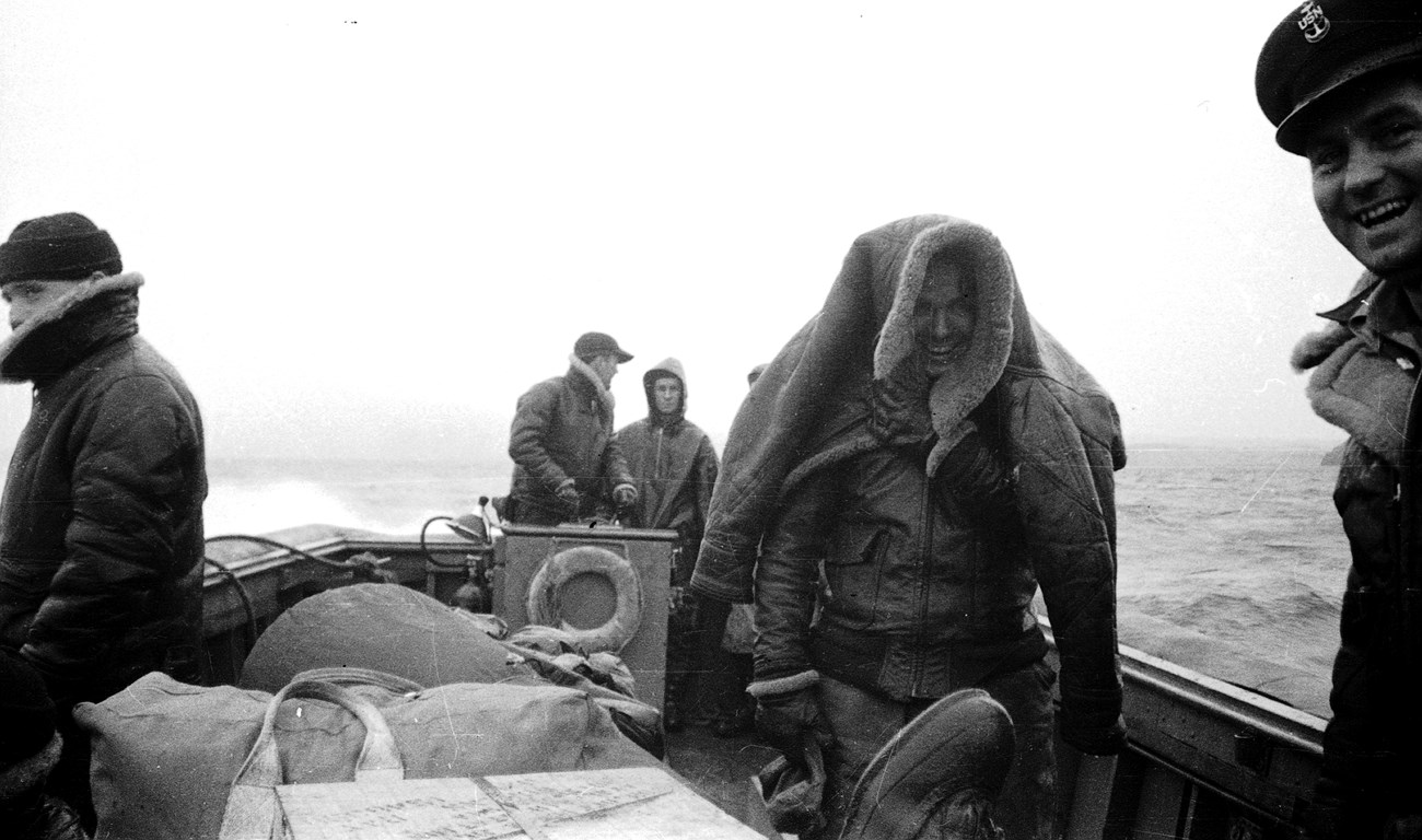 Black and white photo of five men in winter gear and luggage in boat on water.
