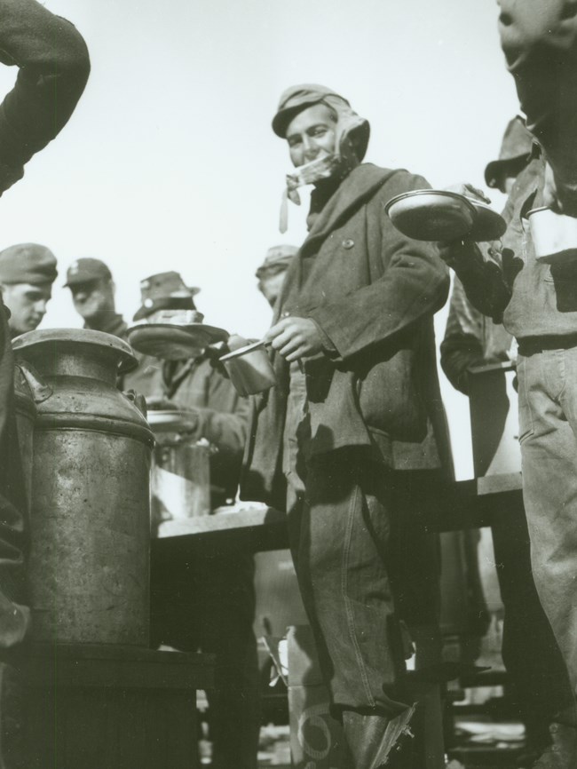 Black and white photo of a young man holding a tin cup and plate; he stands in line with others next to a metal milk can.
