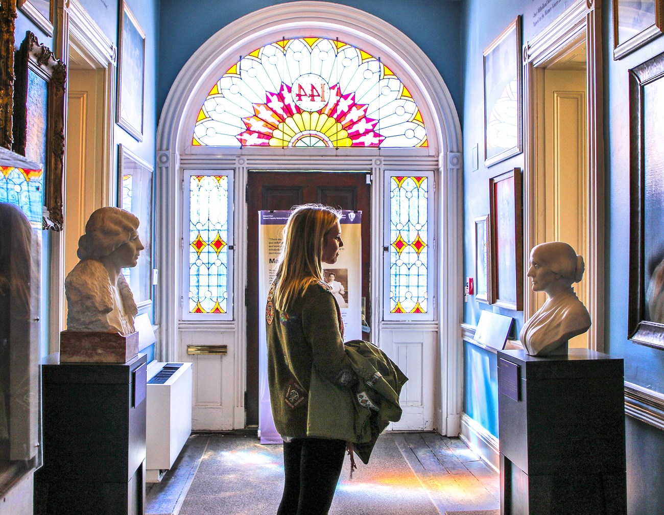 A woman standing between two sculptured busts of suffragists, looking at one. A stained glass window lies above the door in the background, and paintings are on the walls.