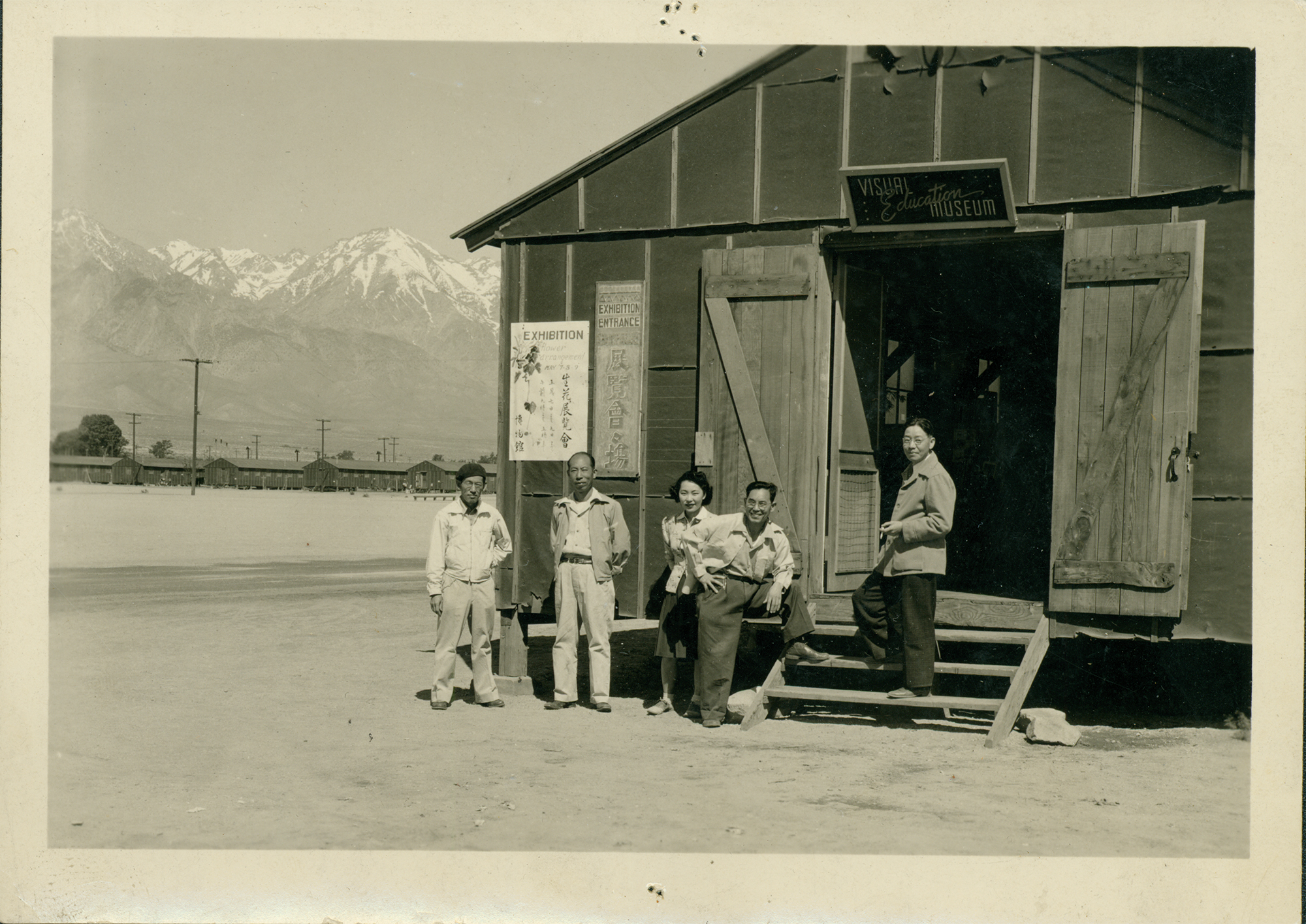 Five individuals standing outside a wooden building with large doors open and two steps made from wooden boards leading into the entrance. A sparce desert landscape leads to snowcapped mountains in the background.