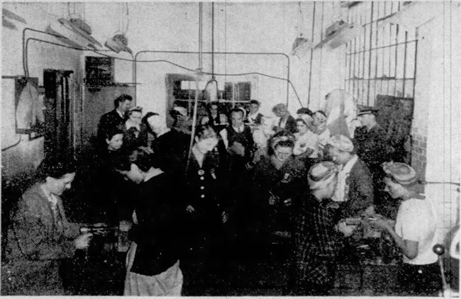 Black and white grainy photograph of groups of women standing over devices on tables. Three men stand in the crowd.