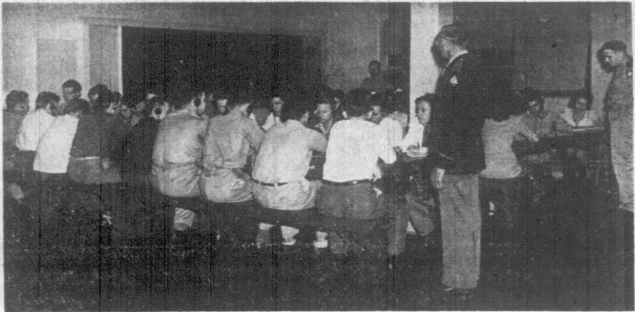 Black and white photo of men seated at a long table with their backs to the camera wearing headphones. A man in uniform stands at one end of the table