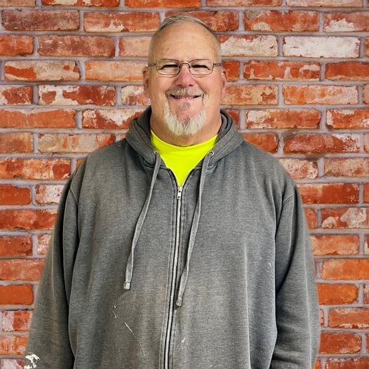 Fredericksburg and Spotsylvania volunteer John Schmiedeke facing the camera and standing in front of a red brick wall. He wears a medium grey zip up hooded sweatshirt over a bright lime green t-shirt.