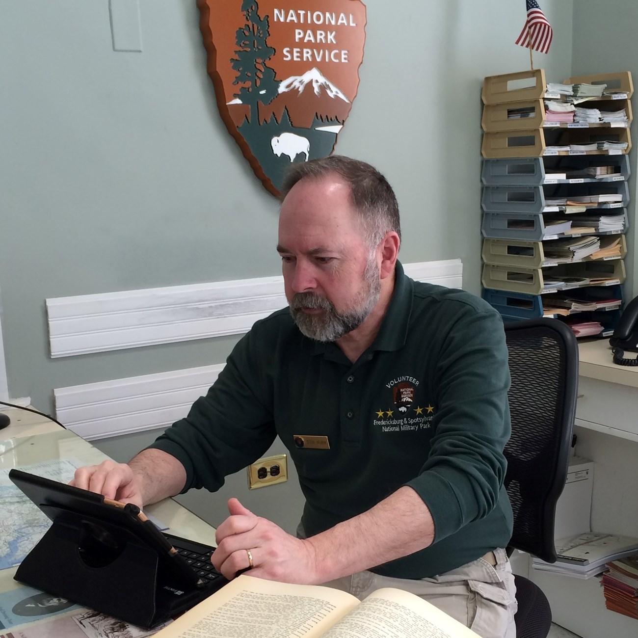 Fredericksburg and Spotsylvania National Military Park volunteer Steve Morin, seated at the Fredericksburg Battlefield Visitor Center front desk. He is doing research work with a laptop and open book on the desk in front of him.
