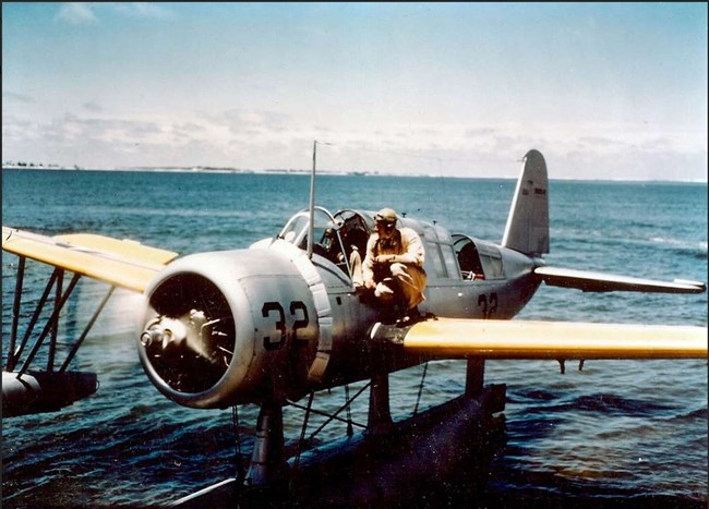 Photo of a silver propeller plane with yellow wings with crouching man on the wing