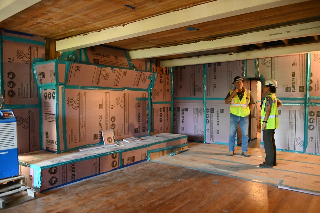 Two people in construction hardhats and reflective vests converse inside a room lined with protective boards.