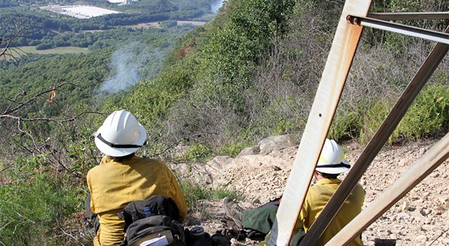 Two wildland firefighters on a hillside watch a plume of smoke on the forested landscape below them.