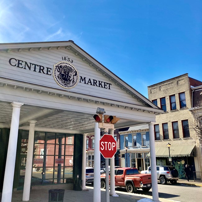 Front of white, columned entrance to building with front portico sign reading "Centre Market" with a seal in between the words and date '1853'. At right, city street with brick buildings.