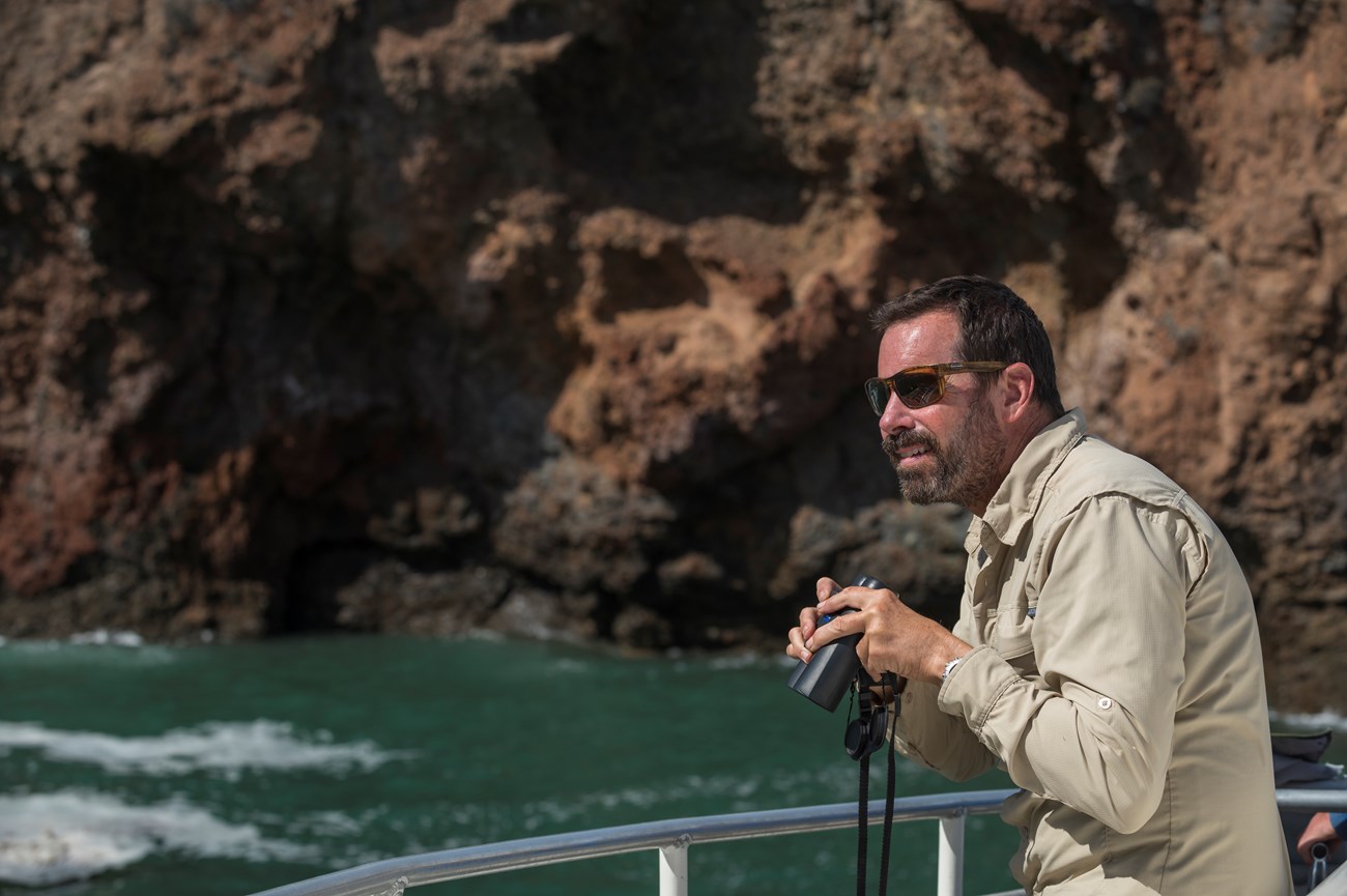 Marine Ecologist, Steve Whitaker holds a pair of binoculars aboard a vessel close to shore. Looking out to viewer's left.