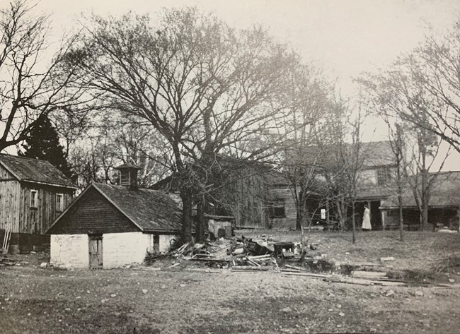 Black and white photo of a rural landscape. An ice house and chicken house are in the foreground while a two story frame house is in the background.