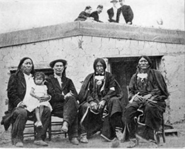 Photo of Arapaho Chief Little Raven sitting next to William Bent with two sons and granddaughter in front of adobe structure