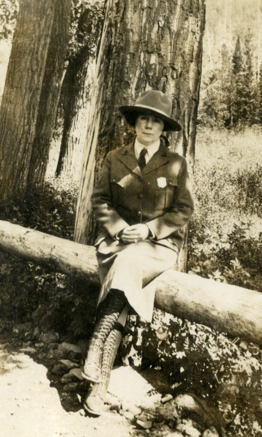 Irene Wisdom sits on a log posing in her NPS uniform skirt, shirt, tie, jacket, and Stetson hat.