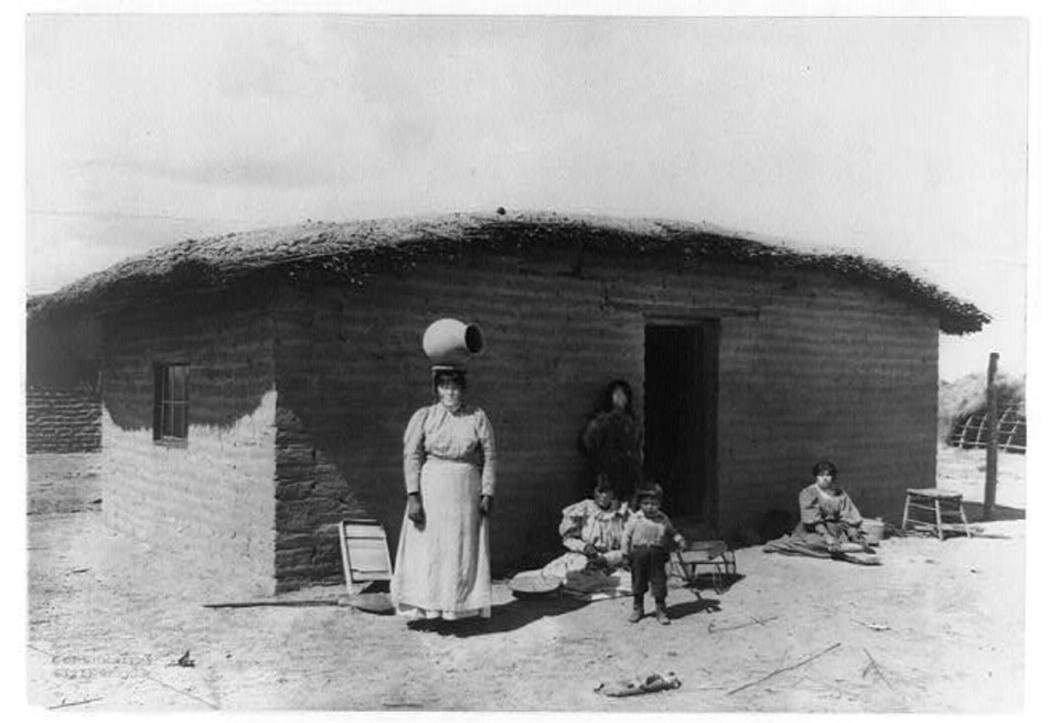 O'odham woman stands outside of adobe house with olla on her head. Four children are posed in the background.
