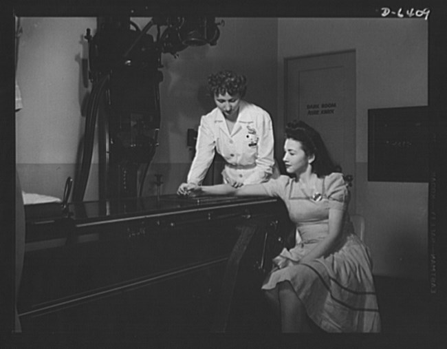 Black and white photo of a uniformed female nurse positioning a woman’s right wrist on an X-ray machine. The woman is seated next to the machine with her right arm propped up onto it.