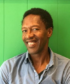 a black man smiling and wearing a gray-blue shirt in front of a green wall