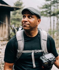 a black man wearing a gray cap, backpack, and carrying a large camera