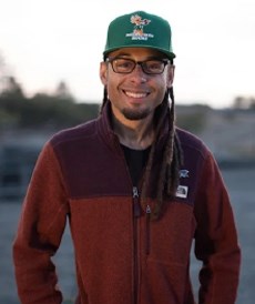 a black man wearing glasses, a green cap, and red sweater with his hair in dreadlocks
