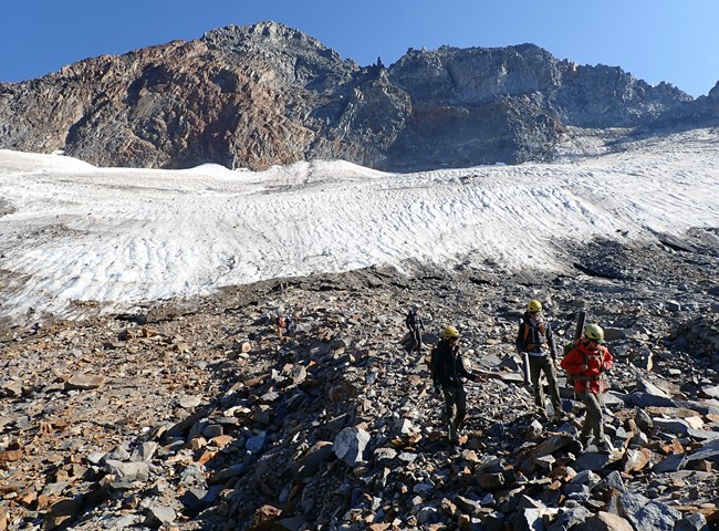 3 people wearing helmets hike across the talus in front of a receding glacier in Yosemite National Park. The mountain in the background is jagged and rocky and it is a clear blue day.