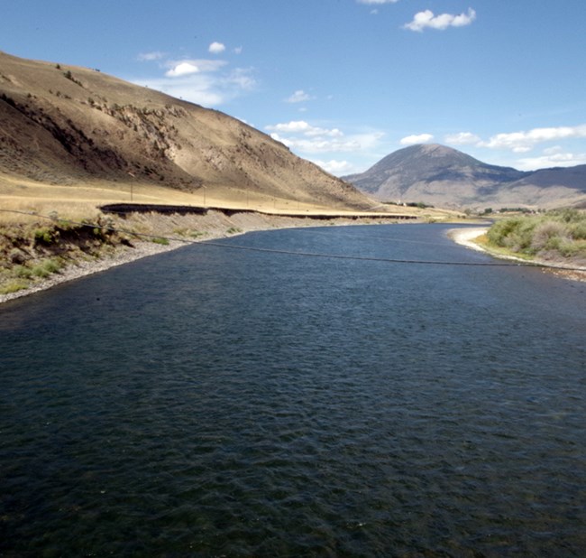 A flowing river lined by grass-covered hills winding away in the distance towards a mountain.