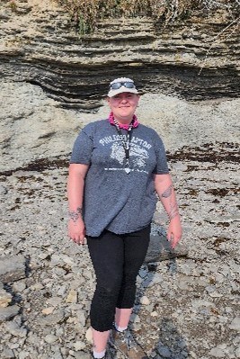 A woman standing in front of a vertical rock outcrop with slightly curved beds of varying thickness and resistance to erosion.