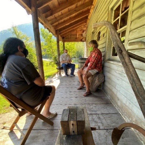 Three people wearing masks sit on outdoor heritage furniture on historic wooden porch