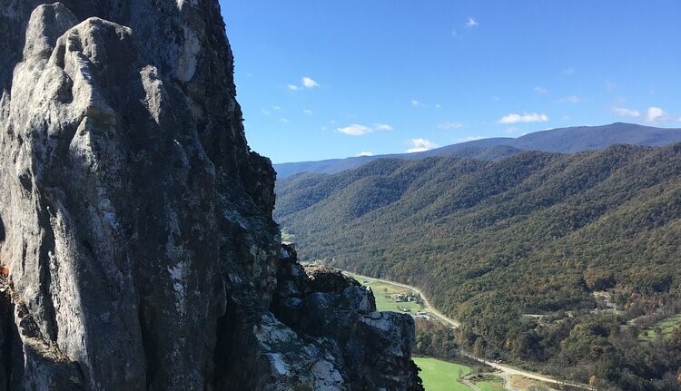 View from mountain overlook. Dramatic craggy rock of peak at left. Looking downward there are small buildings in green valley in distance. Hills and blue sky above in background