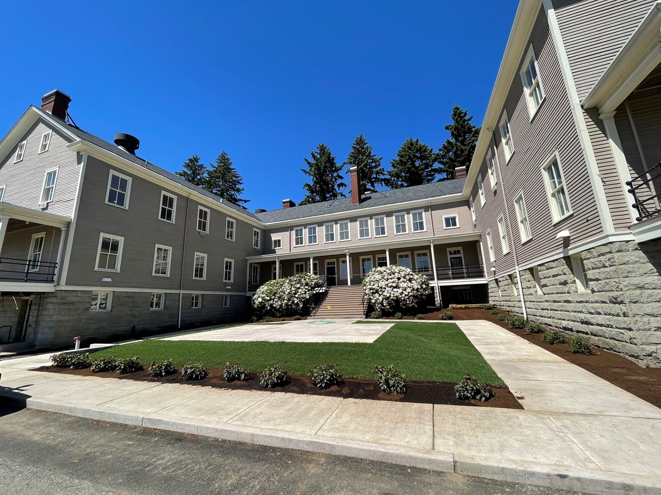 Building 993, a grey building with cream colored trim. The structure is three stories, roughly U-shaped with squared angles, and has a large porch on the central section. A courtyard of shrubs, grass, and pavers is in the foreground.]