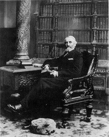 older man sitting in a chair holding a book and he is in front of a large bookcase