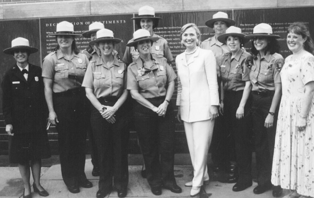 Nine women in NPS uniforms and one in a dress stand in a line posing with Hillary Rodham  Clinton who is wearing a pantsuit.