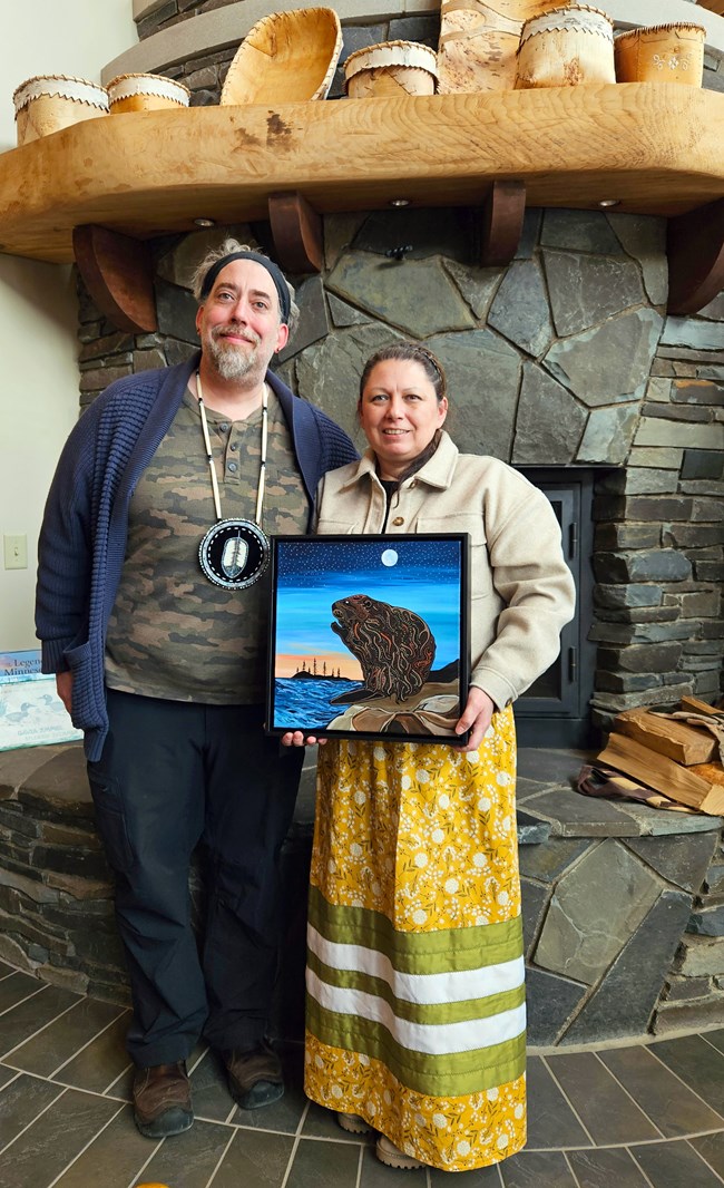Two people standing in front of a stone fireplace with birch baskets on the mantle. One person wears a yellow ribbon skirt and holds a painting of a beaver.