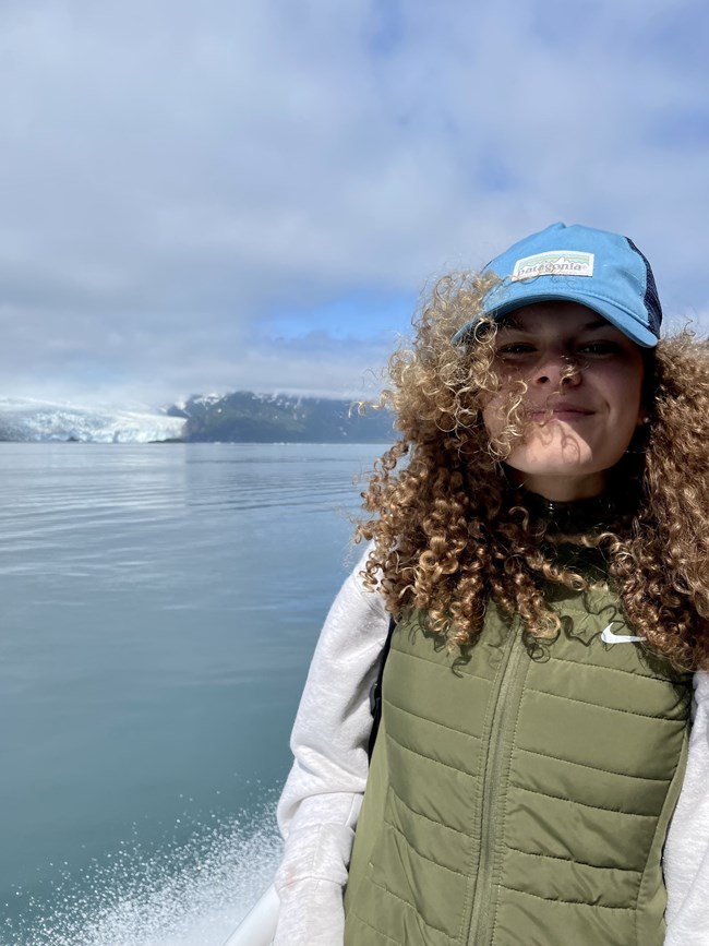 A girl with her hair blowing in the wind with a cap on and glaciers and blue water in the background.