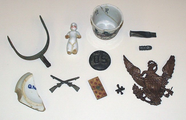 Army artifacts are displayed, including a metal saddle stirrup, a small white china doll, the base of a white ceramic cup marked with the letters “QM” in blue, a bronze insignia in the shape of crossed rifles, a bone domino, and a metal jack.