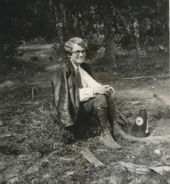 Herma Albertson sits on the ground.  She is wearing a leather coat draped over her shoulders, a long sleeved top and knee boots.