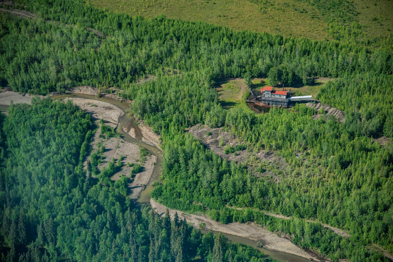 an aerial view of a winding river in a remote forested area with a mining site and building along the bank