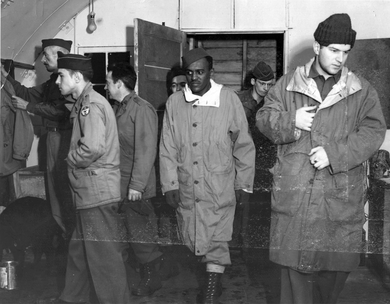 black and white photo of seven men walking into a quonset hut, wearing winter outer clothes.