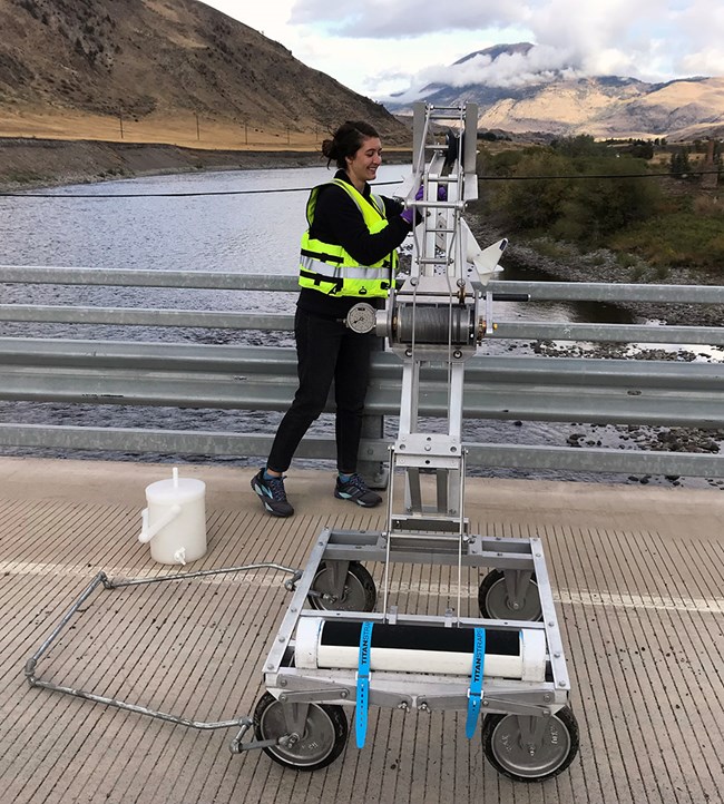 A scientist operating a wheeled crane on a bridge that collects water samples in the river below.