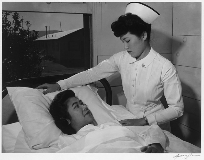 A young East Asian woman in a white nurse’s uniform adjusts the pillow of another East Asian woman lying in a bed.