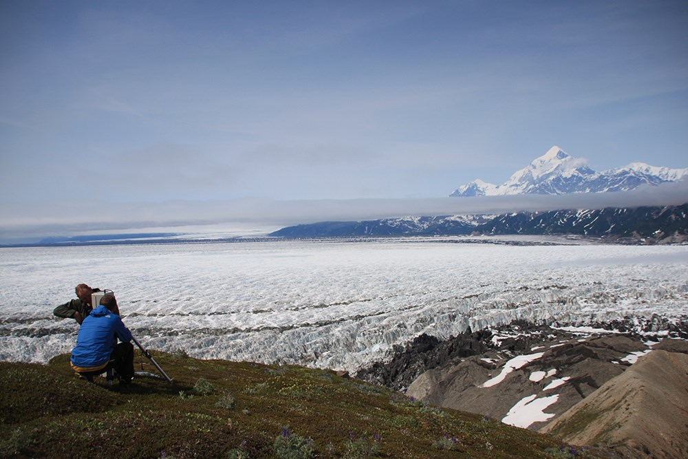 Two researchers install a camera on a tripod overlooking a large glacier.